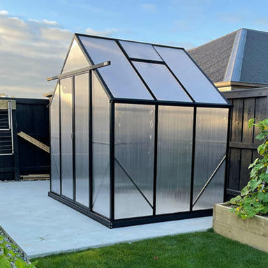 2.6m x 2m polycarbonate greenhouse with black frame sitting on a concrete pad with vegetable gardens & grass.