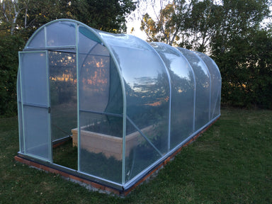 Duratough Tunnel House with open door showing raised garden bed inside, covered with shade cloth. Tunnel house is sitting on bricks on a grass base with trees in background.