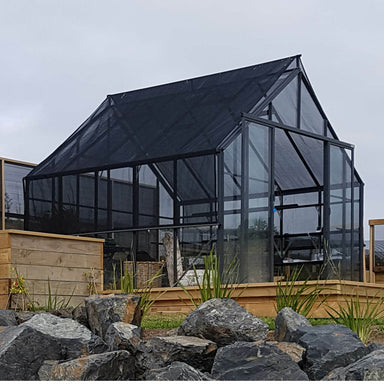 Black framed double door glasshouse with Shade Cover Kit installed, sitting on a wooden deck with chairs inside, gardens & decking around and rocks in the foreground.
