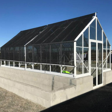 White framed double door glasshouse with Shade Cover kit installed, sitting on a block base with building pebbles around and blue sky in the background.