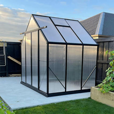 2.6m x 2m polycarbonate greenhouse with black frame sitting on a concrete pad with vegetable gardens & grass. 