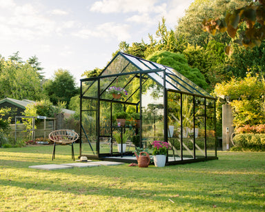 2.6m x 3.2m Black famed glasshouse in lush green backyard with white pebble base and potted flowers inside.