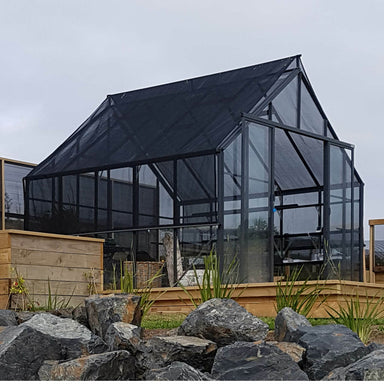 Black framed double door glasshouse with Shade Cover Kit installed, sitting on a wooden deck with chairs inside, gardens & decking around and rocks in the foreground. 