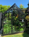 Black framed, single door Glasshouse with shade cover kit over the roof, large green plants and vegetables inside glasshouse with large lush gardens around and blue sky. 