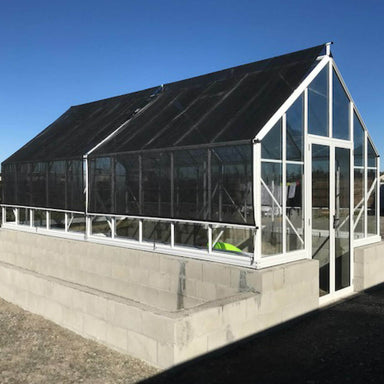 White framed double door glasshouse with Shade Cover kit installed, sitting on a block base with building pebbles around and blue sky in the background. 
